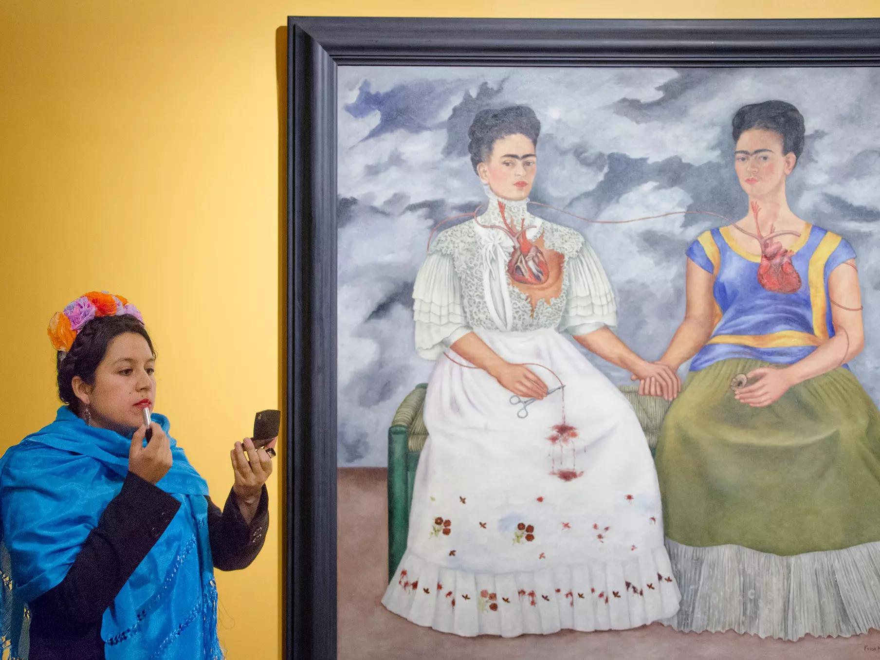 Fotografía de persona caracterizada de Frida Kahlo pintándose los labios a lado de la pintura Las Dos Fridas